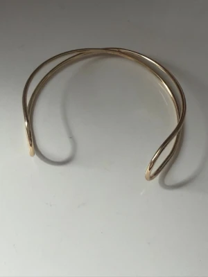 Guldig minimalistisk armring - Säljer en stilren och öppen armring i guldtonad metall. Armbandet har en modern, böjd design med dubbla tunna band som korsar varandra. Perfekt för dig som gillar enkel men unik stil. Passar att bära ensam eller tillsammans med andra smycken.