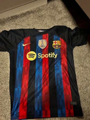 Barcelona matchtröja  - Säljer en FC Barcelona matchtröja från Nike med klassiska blå och röda ränder. Tröjan har Spotify-logga på bröstet, FIFA-märke och klubbmärke. Tillverkad i lätt och ventilerande material, perfekt för fotboll eller träning. UNHCR-logga på ryggen.