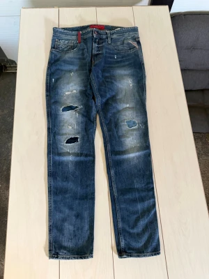 Replay jeans med Blå slitna detaljer & hål - Säljer ett par blå jeans med slitningar och hål på benen. Jeansen har fem fickor, normal midja och raka ben. Snyggt tvättade detaljer och en röd etikett vid fickan ger extra stilpoäng. Perfekta för en avslappnad look.