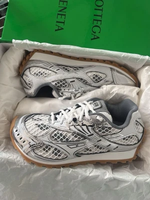 Vita sneakers från Bottega Veneta - Snygga vita sneakers från Bottega Veneta med coolt ormmönster i silver och svart. Skorna har vita snören, grå insida och en chunky gummisula i beige. Perfekt för dig som vill sticka ut med en unik och trendig look.