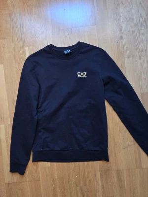 Svart sweatshirt från EA7 Emporio Armani - Stilren svart sweatshirt från EA7 Emporio Armani med rund halsringning och diskret logga på bröstet. Tröjan har långa ärmar, ribbade muddar och en gulddetalj i nacken. Perfekt för en avslappnad och trendig look.