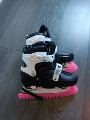 Svarta och vita skridskor med skydd - Säljer ett par svarta och vita skridskor med rosa skydd på skenorna. Skridskorna har justerbara spännen och är tillverkade i plast och syntetmaterial. Perfekta för isbanan eller hockeyträningen. Märket Graf syns på sidan. Använda en säsong ställbara från 34 till 37
