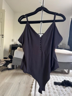 Svart body med knappar framtill - Säljer en svart body med smala axelband och v-ringning. Framtill finns dekorativa knappar som ger en snygg detalj. Materialet är mjukt och stretchigt, vilket gör den bekväm att bära. Perfekt att styla till olika outfits.