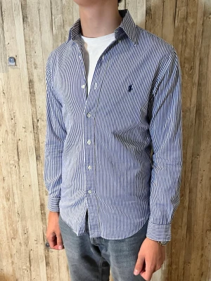 Blårandig skjorta med broderad logga - Snygg skjorta med smala blå och vita ränder och klassisk krage. Skjortan har långa ärmar, knäppning framtill och en liten broderad logga på bröstet. Perfekt till jeans eller chinos för en clean look.