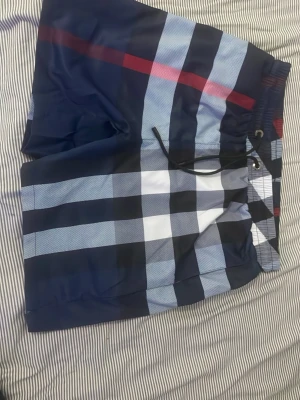 Rutiga badbyxor i blått och vitt - Helt nya snygga badbyxor med rutigt mönster i marinblått, ljusblått, vitt och en röd detalj. Klassisk modell med dragsko i midjan och meshfoder. Perfekta för stranden eller poolen, ger en cool och avslappnad look.