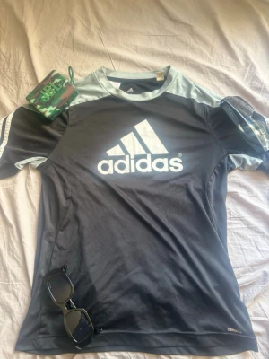Svart sporttröja från Adidas - Snygg svart sporttröja från Adidas med stora vita loggan på bröstet och ljusgrå detaljer vid axlar och hals. Tillverkad i ett lätt och snabbtorkande material, perfekt för träning eller sportaktiviteter. Klassisk passform och korta ärmar. Pris kan diskuteras.