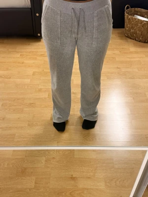 Grå juciy couture byxor - Säljer ett par grå mjukisbyxor med bred resår i midjan och dragsko. Byxorna har två stora fickor fram och ett diskret broderat märke på ena fickan. Sköna och avslappnade med rak passform, perfekta för chill dagar hemma. Byxorna är i storlek xs men dom har varit uppsydda för dom va för långa. Passar bra om du är kortare än 165cm💕