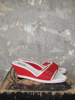 Röda kilklackssandaler från Lacoste - Röda sandaler med kilklack från Lacoste. De har vita detaljer och den klassiska krokodilloggan på sidan. Öppen tå och bred rem över foten. 