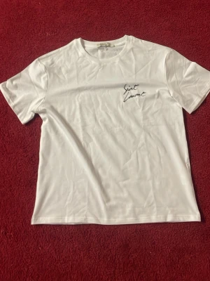 Vit Saint Laurent t-shirt - Vit t-shirt från Saint Laurent med svart logotyptryck både fram och bak. Klassisk passform med rund hals och korta ärmar. Perfekt för dig som gillar stilrena och trendiga plagg med exklusiv känsla.