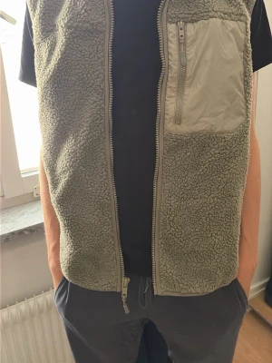 Olivgrön fleeceväst med dragkedja - Mysig olivgrön fleeceväst med hel dragkedja framtill och en bröstficka i nylon med dragkedja. Västen har en enkel, rak passform och är perfekt för lager-på-lager. Materialet är mjukt teddyfleece som håller värmen och ger en avslappnad look. Lappen sitter kvar