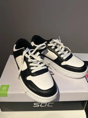Svarta och vita sneakers från SOC - Snygga sneakers i svart och vitt från SOC. Skorna har klassisk låg profil, vita snören och perforerad tå för extra ventilation. Ovandelen är i konstläder med kontrasterande paneler och en tjock vit sula som ger en sportig look.