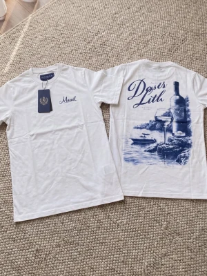 Vit t-shirt med blått tryck - Vit t-shirt med blått tryck på ryggen som föreställer vinflaska, glas och naturmotiv. Framsidan har en liten blå text vid bröstet. Klassisk passform och rund halsringning. Perfekt för en stilren och avslappnad look.