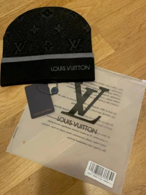 Svart mössa från Loois Voitton - Snygg svart mössa från Loois Voitton med klassiskt monogrammönster och grå rand. Märkets logga är broderad framtill. Mössan är tillverkad i mjukt material som känns lyxigt och bekvämt. Perfekt accessoar för dig som vill ha en exklusiv touch på din outfit. Priset kan sänkas 