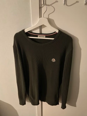 Mörkgrön stickad tröja Moncler - Snygg mörkgrön stickad tröja från Moncler med rund halsringning och diskret Moncler-logga på bröstet. Tröjan har lång ärm och ribbstickad struktur som ger en clean och stilren look. Perfekt för dig som gillar klassiska plagg med exklusiv känsla.