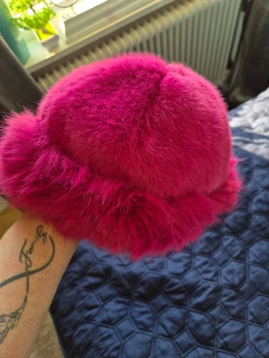 Fuskpäls rosa bucket hat - Säljer en fluffig bucket hat i starkt rosa fuskpäls. Hatten har en rundad form och bred kant, perfekt för dig som vill sticka ut med en färgglad accessoar. Materialet är mjukt och ger en lyxig känsla. Märke saknas.