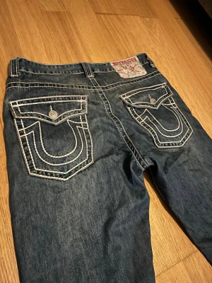 True Religion blå jeans med kontrastsömmar - Säljer ett par blå jeans från True Religion med tydliga vita kontrastsömmar och stora ficklock bak. Klassisk femficksmodell med logga på bakfickan och metallknappar. Tillverkade i denim med en avslappnad passform och detaljerade sömmar.
