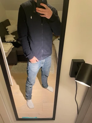 Svart dunväst från Polo Ralph Lauren - Snygg svart dunväst från Polo Ralph Lauren med diskret broderad logga på bröstet. Västen har hög krage, dragkedja framtill och quiltad design. Perfekt att slänga över en hoodie eller tröja för extra värme och stil. 