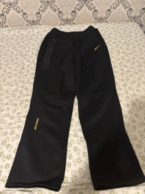 Svarta NOCTA mjukisbyxor från Nike - Säljer ett par svarta mjukisbyxor från Nike NOCTA-kollektionen. Byxorna har raka ben, dragsko i midjan och gula detaljer med NOCTA-logga och Nike Swoosh. Tillverkade i mjukt material som passar perfekt för chill dagar. Snygg streetwear vibe med broderade detaljer.