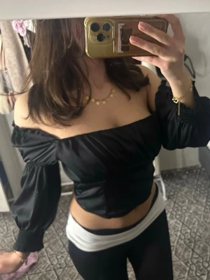 Svart offshoulder topp i satin - Trendig svart offshoulder topp i glansigt satinliknande material. Toppen har långa puffärmar med resår vid ärmslut och sitter tight över bysten och midjan för en snygg siluett. Perfekt för dig som vill ha en statement piece till din outfit.