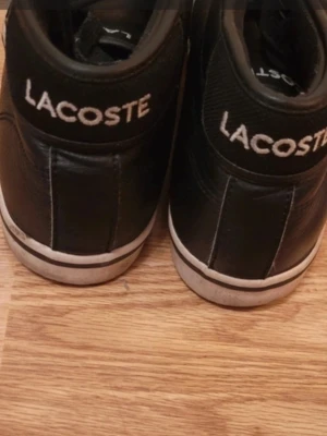 Svarta Lacoste sneakers i läder - Snygga svarta sneakers från Lacoste i äkta läder med klassisk vit sula och broderad grön krokodil på sidan. Skorna har svarta skosnören och Lacoste-logga broderad bak på hälen. Perfekta för en clean och stilren look.