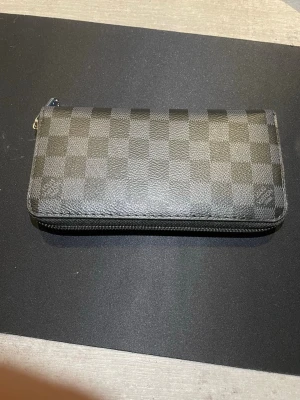  rutmönstrad plånbok - Snygg plånbok i svart och grått med det klassiska rutmönstret Damier Graphite. Tillverkad i tåligt canvasmaterial och har dragkedja runtom. Perfekt för dig som gillar exklusiva accessoarer med stilren design.