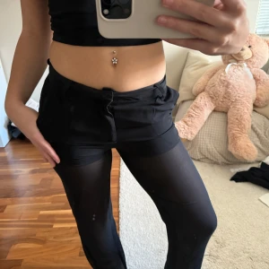 Svarta kostymbyxor - Säljer ett par svarta kostymbyxor med transparent mesh på benen och knappdetalj i midjan. De har en tight passform upp till och nertill raka. låg midja, perfekta för en trendig och edgy look. Materialet är stretchigt och bekvämt.