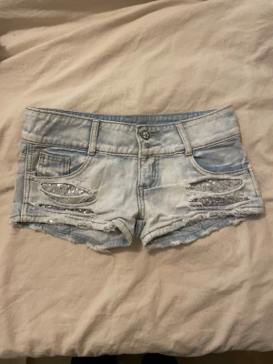  jeansshorts  - Säljer ett par ljusblå jeansshorts med råa kanter och slitningar framtill. Shortsen har silverfärgade paljettdetaljer i slitningarna och klassiska fem fickor. Perfekta för en avslappnad och trendig sommarlook.