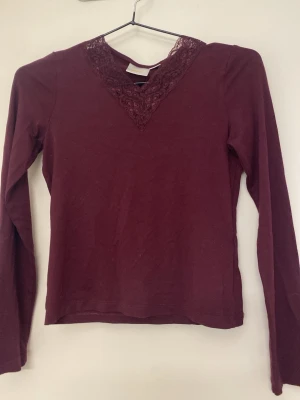 Vinröd långärmad topp med spets - Vinröd långärmad topp med v-ringning och dekorativ spetsdetalj vid halsen. Tunn och mjuk, perfekt att styla med jeans eller kjol. Enkel men ändå elegant design som passar till många olika looks.