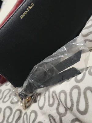 Svart plånbok från Anna Field - Stilren svart plånbok från Anna Field i slätt konstläder. Plånboken har guldfärgad logga framtill och dragkedja med matchande detaljer. Perfekt för dig som gillar enkel och klassisk design.