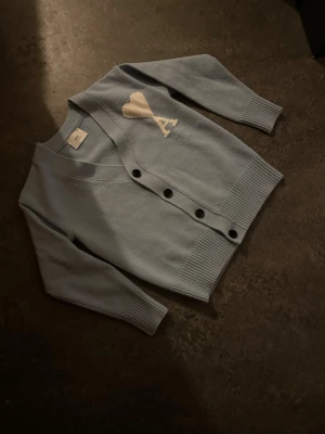 Ljusblå cardigan från Ami Paris - Supersnygg ljusblå cardigan från Ami Paris med svart knappstängning och ribbade muddar. Tröjan har ett vitt hjärta med bokstaven A på bröstet och är gjord i mjukt stickat material. Perfekt för dig som gillar stilrena och trendiga plagg.