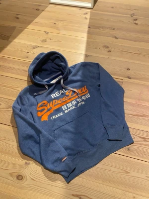 Blå Superdry hoodie med tryck - Snygg blå hoodie från Superdry med stort orange och vitt tryck på bröstet. Klassisk känguruficka framtill, dragsko i huvan och ribbade muddar. Perfekt för en avslappnad streetstyle-look. Mjuk bomullsblandning för extra komfort.tveka inte vid fråger!😁