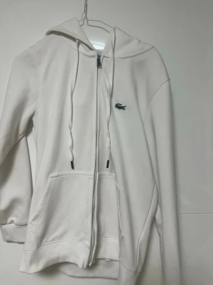Vit zip hoodie från Lacoste - Snygg vit hoodie från Lacoste med dragkedja framtill, klassisk krokodil-logga på bröstet och stora fickor. Hoodien har justerbar huva med snören och ribbade muddar vid ärmar och nederkant. Tillverkad i mjukt bomullsmaterial för skön känsla.
