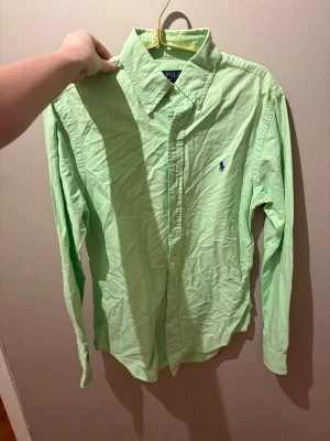 Grön skjorta från Polo Ralph Lauren - Snygg ljusgrön skjorta från Polo Ralph Lauren med klassisk button-down krage och blå broderad logga på bröstet. Skjortan har långa ärmar och knäppning framtill. Perfekt för en clean och stilren look.