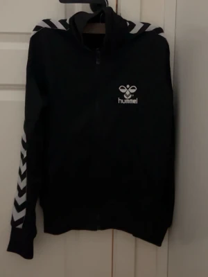Svart Hummel ziptröja med chevrons - Säljer en svart ziptröja från Hummel med vita chevronmönster på ärmarna och axlarna. Tröjan har dragkedja framtill och Hummel-logga på bröstet. Perfekt till träning eller chill. Klassisk sportig look.