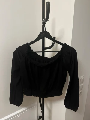 Svart croppad off shoulder topp med puffärmar - Trendig svart croppad off shoulder topp med långa puffärmar och smockdetaljer vid halsringning och midja. Tillverkad i mjukt material med 99% polyester och 1% elastan för stretchig passform. Perfekt för att lyfta vilken outfit som helst.