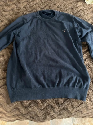 Mörkblå sweatshirt från GANT - Klassisk mörkblå sweatshirt från GANT med rund halsringning och diskret logga broderad på bröstet. Tröjan har ribbade muddar vid ärmslut och nederkant. Perfekt för en avslappnad och stilren look.