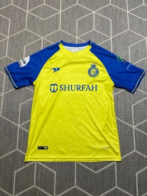 Al Nassr matchtröja - Snygg gul och blå matchtröja från Al Nassr FC med klubbmärke på bröstet och SHURFAH-tryck på framsidan. Tröjan har korta ärmar, rund hals och är tillverkad i ett lätt och ventilerande funktionsmaterial. Perfekt för fotbollsträning eller att bära till vardags.