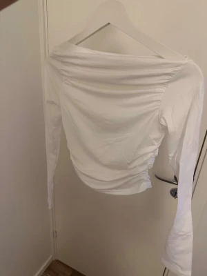 Vit offshoulder topp med rynk - Trendig vit offshoulder topp med långa ärmar och snygga rynkdetaljer på sidorna. Toppen har en croppad passform och är tillverkad i ett mjukt bomullsliknande material. Perfekt för dig som vill ha en stilren men ändå unik look.