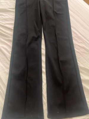 Svarta vida kostymbyxor - Säljer ett par svarta kostymbyxor med pressveck och utsvängda ben. Byxorna har en klassisk look och är perfekta för en stilren outfit. Materialet känns mjukt och följsamt, vilket ger en snygg passform.