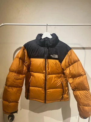 The North Face jacka - Säljer en klassisk dunjacka från The North Face i brunt och svart. Jackan har hög krage, dragkedja framtill och två sidofickor. Puffig modell med logga på bröstet och ryggen. Perfekt för kalla vinterdagar och riktigt snygg streetstil.