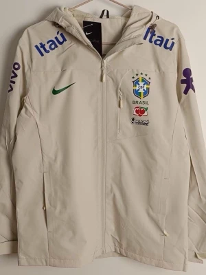 Nike Brasil vindjacka beige - Säljer en beige Nike vindjacka med Brasil-logga och flera coola sponsortryck. Jackan har huva, hel dragkedja framtill och praktiska fickor. Perfekt för dig som gillar sportig stil och vill sticka ut med landslagsvibbar.