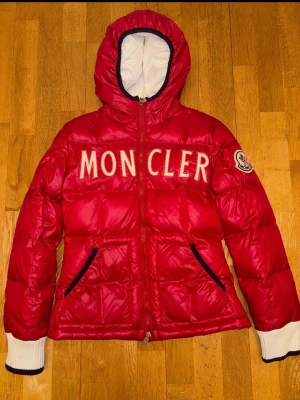 Moncler dunjacka barn – Röd – Strl 12 år / 152 cm - Snygg röd dunjacka från Moncler med stor logga broderad över bröstet och patch på ärmen. Jackan har huva, vita ribbade muddar med svart kant och två fickor med svarta detaljer. Perfekt för kalla dagar och riktigt trendig look.