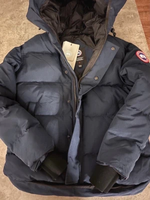 Mörkblå dunjacka från Canada Goose - Snygg mörkblå dunjacka från Canada Goose med huva och klassisk logga på ärmen. Jackan har stora fickor framtill, kraftig dragkedja och muddar vid ärmsluten. Vatten tätt.Perfekt för kalla vinterdagar och riktigt varm tack vare dunfyllningen.
