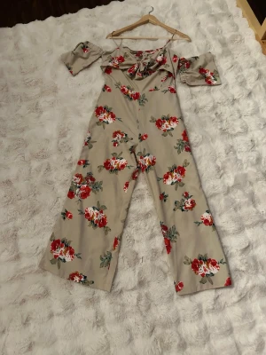 Blommig somrig jumpsuit från Boohoo - Snygg beige jumpsuit från Boohoo med röda och vita blommor. Off shoulder-modell med knytning framtill och vida ben. Perfekt för dig som vill sticka ut med ett färgglatt och trendigt plagg. Tunn och skön kvalitet.