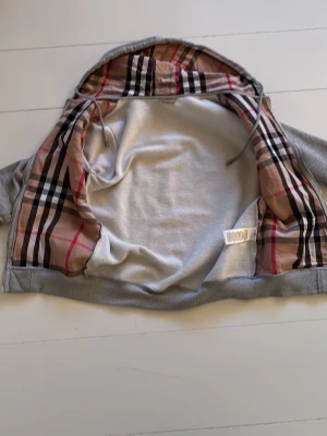 Grå hoodie från Burberry med dragkedja - Pris kan diskuteras. Snygg grå hoodie från Burberry med dragkedja och klassiskt rutigt foder i huvan. Hoodien har två fickor framtill och ribbade muddar vid ärmslut och nederkant. Perfekt för en chill och stilren look. Materialet är mjukt och bekvämt, troligen bomullsblandning.  Mycket bra skick.   Pris kan diskuteras 