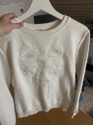 Vit sweatshirt från Kenzo med tiger - Säljer en vit sweatshirt från Kenzo med det ikoniska tigermotivet broderat framtill och texten 'Kenzo Paris'. Tröjan har rund halsringning, ribbade muddar och är i mjukt material. Perfekt för dig som gillar streetwear och vill ha något stilrent men ändå unikt.