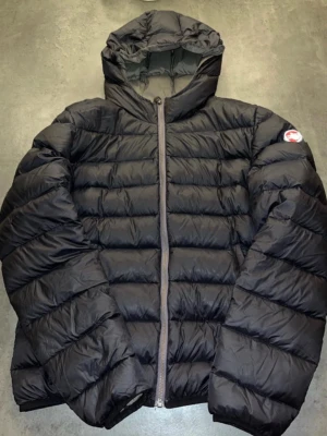 Svart colmar jacka - Snygg svart dunjacka från Colmar med huva och dragkedja framtill. Jackan har ett quiltat mönster och en rund logotyp på ärmen. Perfekt för kalla vinterdagar och riktigt skön att bära.