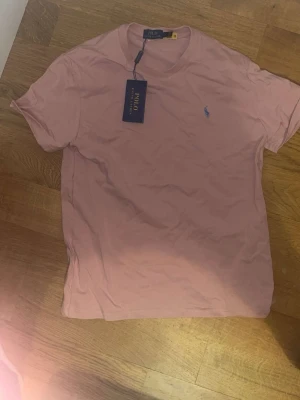 Rosa t-shirt från Polo Ralph Lauren - Snygg rosa t-shirt från Polo Ralph Lauren med klassisk passform och korta ärmar. T-shirten har en liten blå logga broderad på bröstet och är tillverkad i mjuk bomull. Perfekt för en avslappnad och stilren look. 