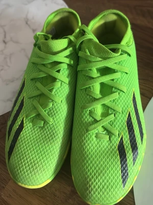 Neongröna Adidas fotbollsskor - Säljer ett par neongröna fotbollsskor från Adidas med klassiska svarta ränder på sidorna. Skorna har snörning och ovandel i syntetmaterial med texturerad yta för bättre bollkontroll. Perfekta för dig som vill sticka ut på planen.