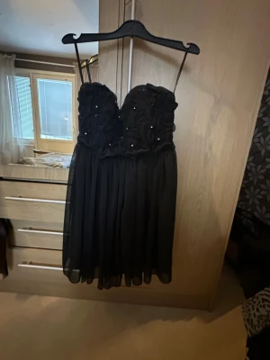 Svart kort klänning med pärlor - Snygg svart kort klänning med tunna axelband och dekorerad överdel med blommiga detaljer och små pärlor. Kjolen är lätt och luftig i chiffongliknande material. Perfekt för dig som vill ha en elegant look med det lilla extra.